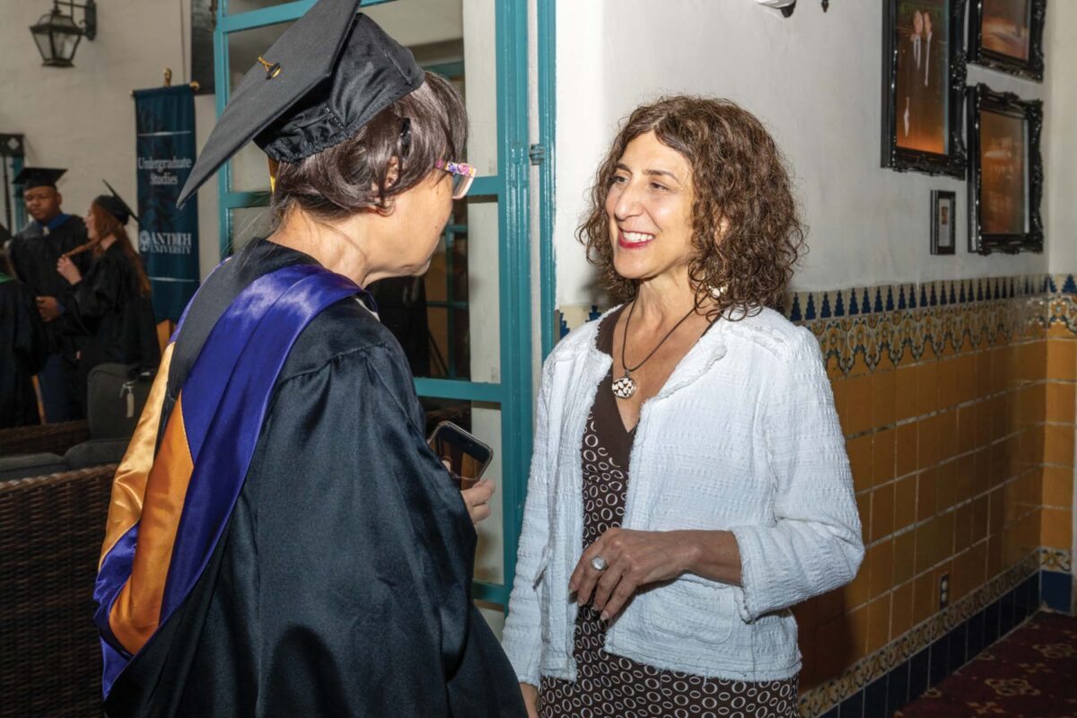 Graduate in cap and gown chats with a smiling woman by a doorway in a school hallway.