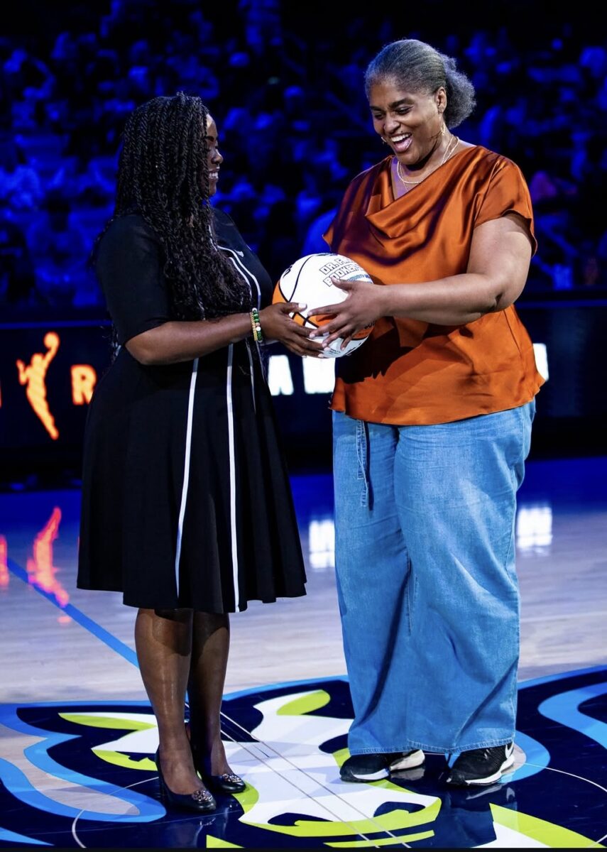 Froswa Booker, wearing a copper blouse and denim skirt, smiles as she presents a commemorative basketball to another woman during a recognition ceremony on a basketball court. Both are smiling and holding the basketball together, with the arena and audience softly blurred in the background.