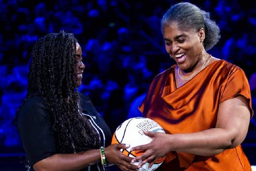 Froswa Booker, wearing a copper blouse and denim skirt, smiles as she presents a commemorative basketball to another woman during a recognition ceremony on a basketball court. Both are smiling and holding the basketball together, with the arena and audience softly blurred in the background.