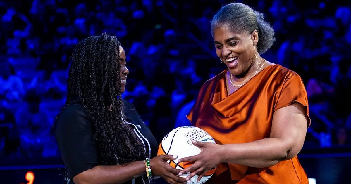 Froswa Booker, wearing a copper blouse and denim skirt, smiles as she presents a commemorative basketball to another woman during a recognition ceremony on a basketball court. Both are smiling and holding the basketball together, with the arena and audience softly blurred in the background.