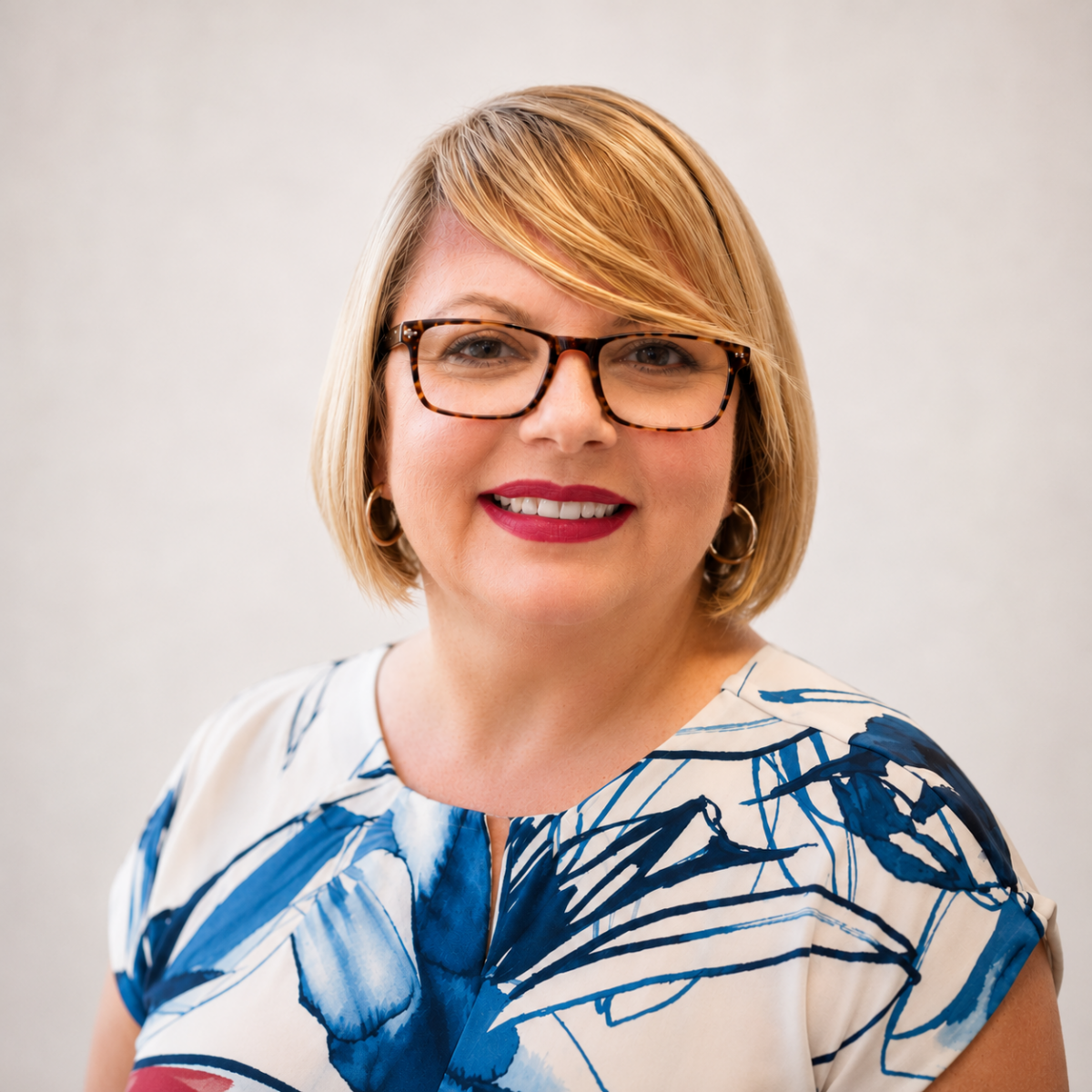 Headshot of Kader Gümüş smiling at the camera. She has a short blonde bob, tortoiseshell glasses, hoop earrings, and a white blouse with a bold blue floral pattern, posed against a softly lit neutral background.