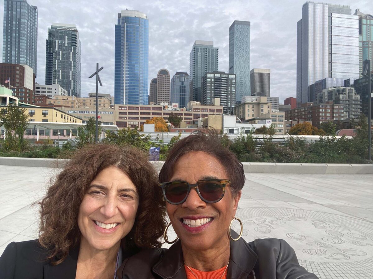 Two smiling adults take a selfie with a city skyline of tall glass buildings behind them.