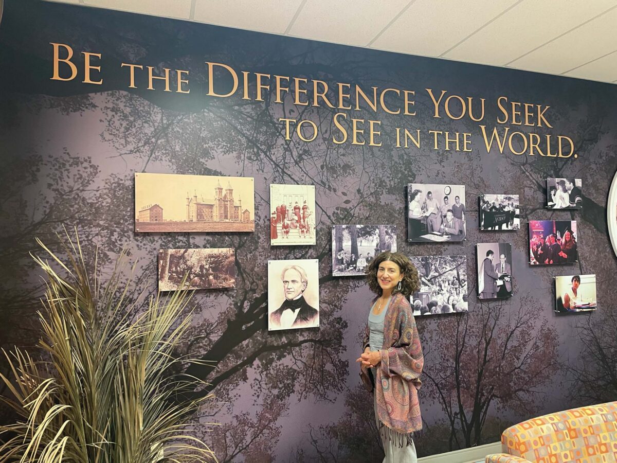 A woman stands in front of a dark wall with a large gold quote, 'Be the Difference You Seek to See in the World,' and vintage photo panels.