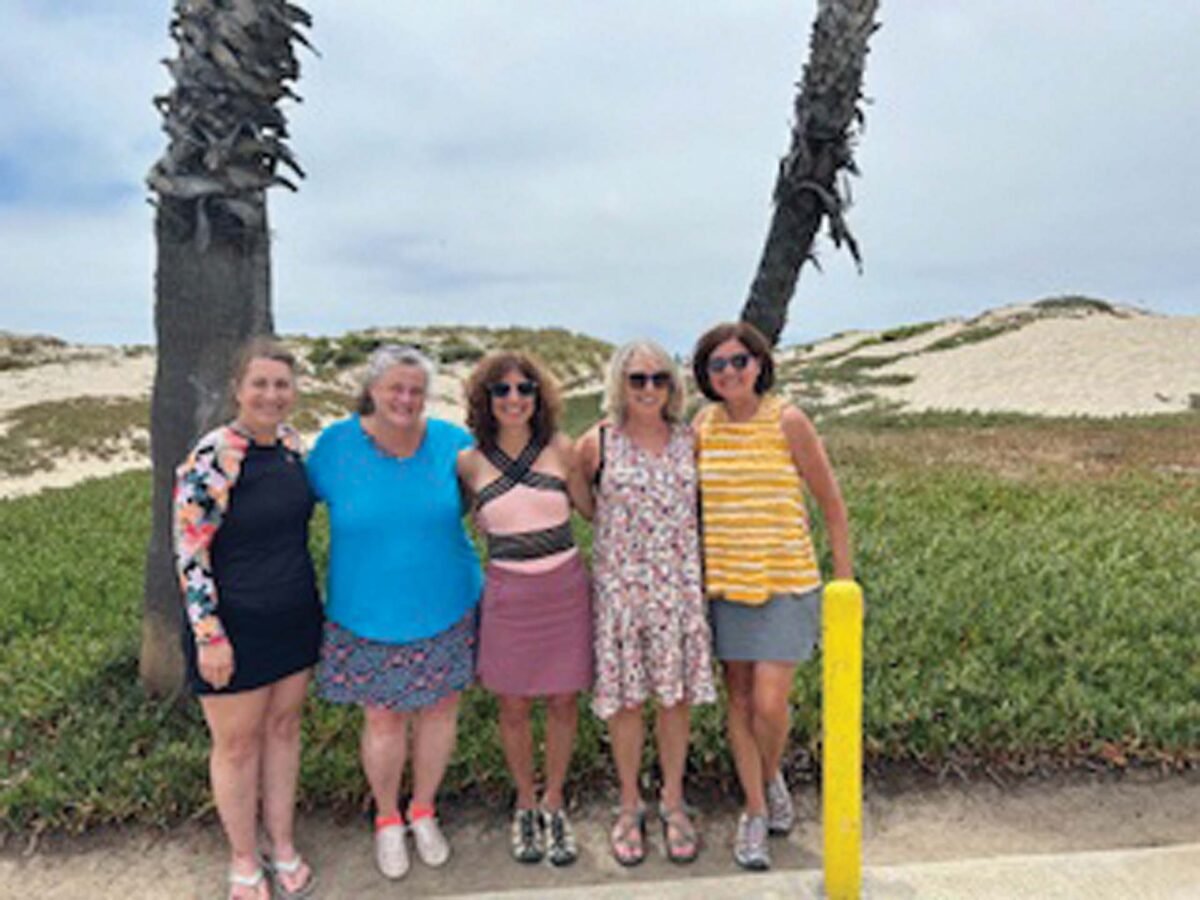 Five women standing together on a sandy path near dunes, smiling at the camera on a sunny day at the beach.