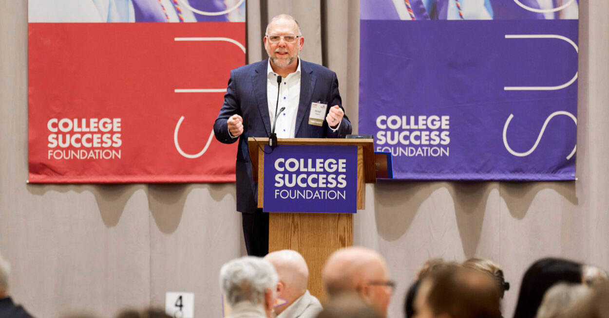 Michael Cheever, wearing a navy suit gives a talk at a podium with College Success Foundation banners in red and purple behind him.
