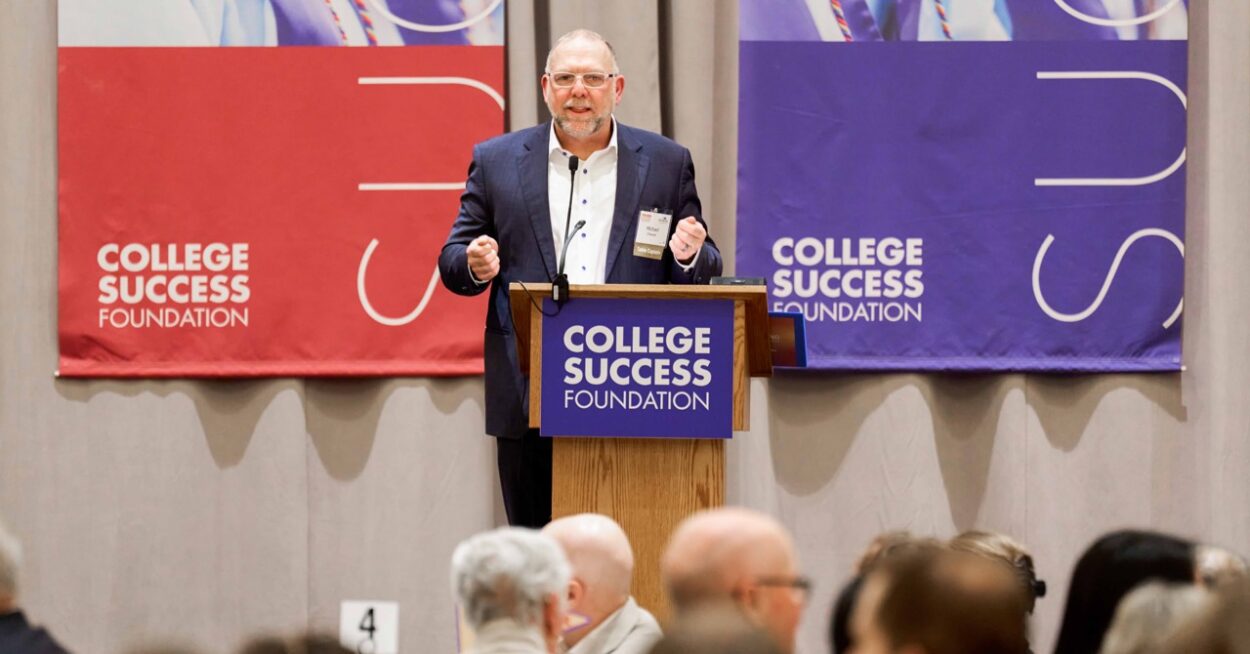 Michael Cheever, wearing a navy suit gives a talk at a podium with College Success Foundation banners in red and purple behind him.