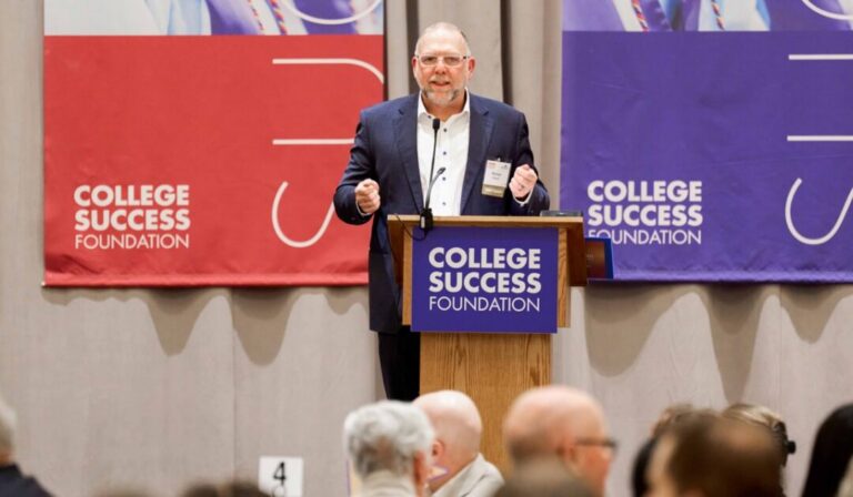 Michael Cheever, wearing a navy suit gives a talk at a podium with College Success Foundation banners in red and purple behind him.