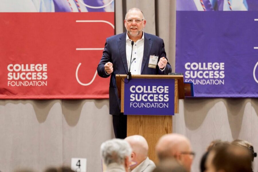 Michael Cheever, wearing a navy suit gives a talk at a podium with College Success Foundation banners in red and purple behind him.