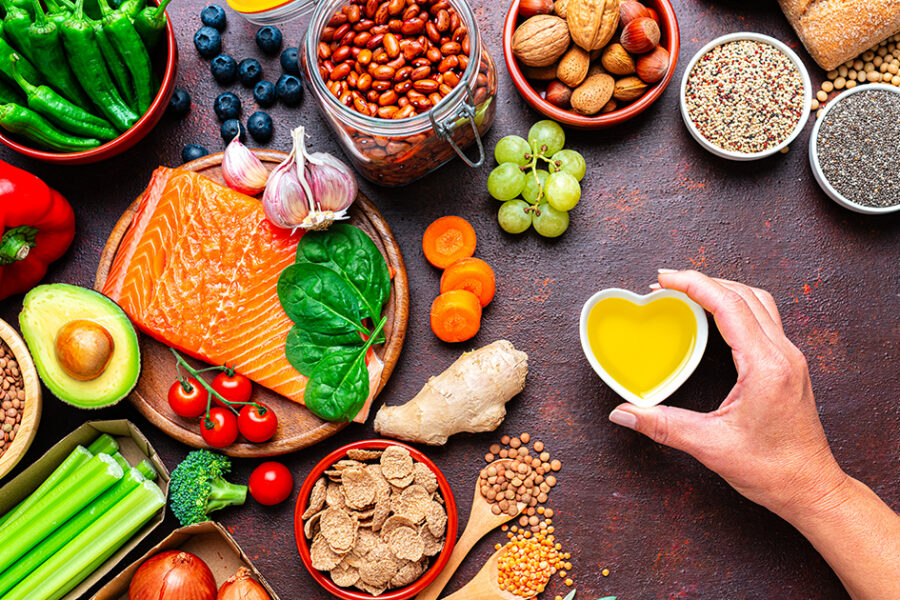 Overhead view of a variety of healthy foods, including salmon, leafy greens, berries, beans, nuts, whole grains, vegetables, avocado, and olive oil, arranged around a hand holding a small heart-shaped bowl of olive oil, representing a heart-healthy, nutrient-rich diet.