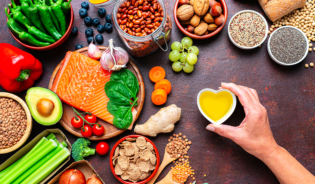 Overhead view of a variety of healthy foods, including salmon, leafy greens, berries, beans, nuts, whole grains, vegetables, avocado, and olive oil, arranged around a hand holding a small heart-shaped bowl of olive oil, representing a heart-healthy, nutrient-rich diet.
