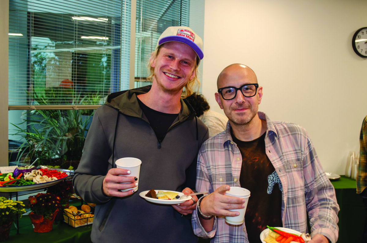 Two men smiling at a table with snacks, holding cups at a casual gathering.
