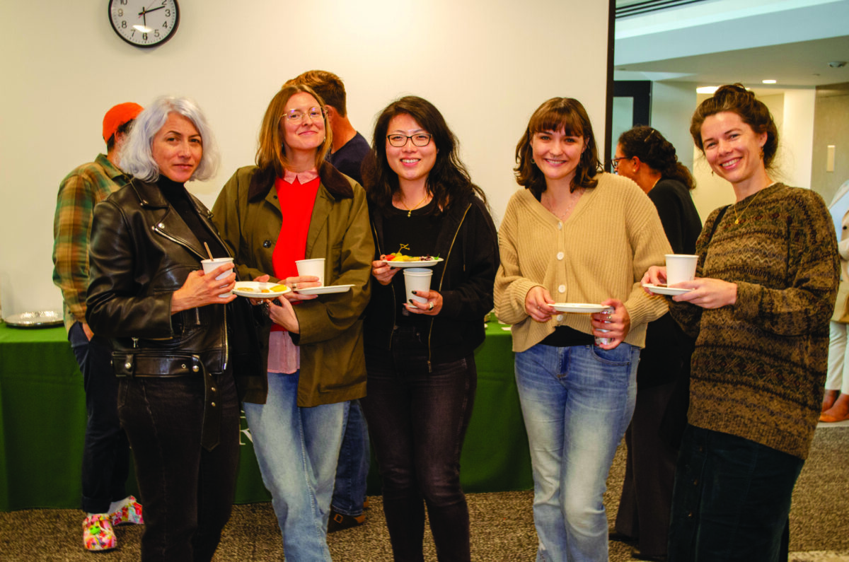 Group of six people standing together at a casual gathering, holding plates and cups, smiling at the camera in a room with a clock on the wall.