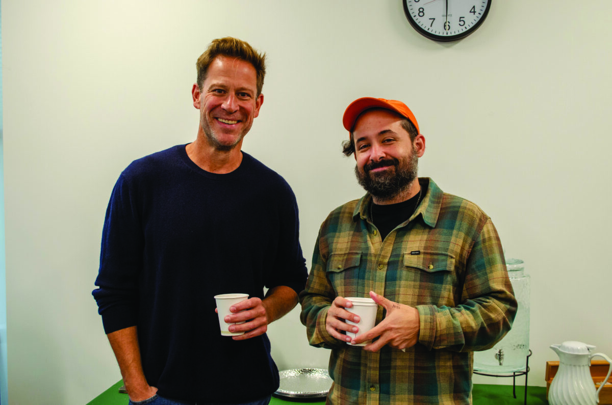 Two men smiling and holding white coffee cups in a casual indoor setting.