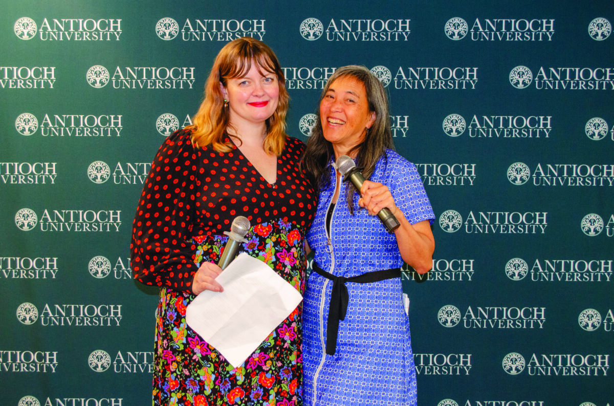 Two smiling women pose for a photo at an Antioch University event, one holding a microphone and papers against a branded backdrop.
