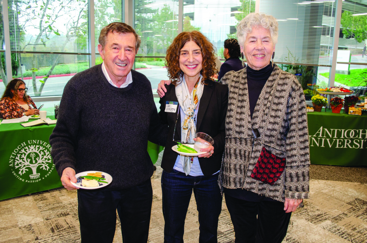 Three adults smiling for a photo at a university event; man on the left holds a plate, center woman wears a scarf and name badge, right woman wears a patterned coat with a red plaid pocket detail.