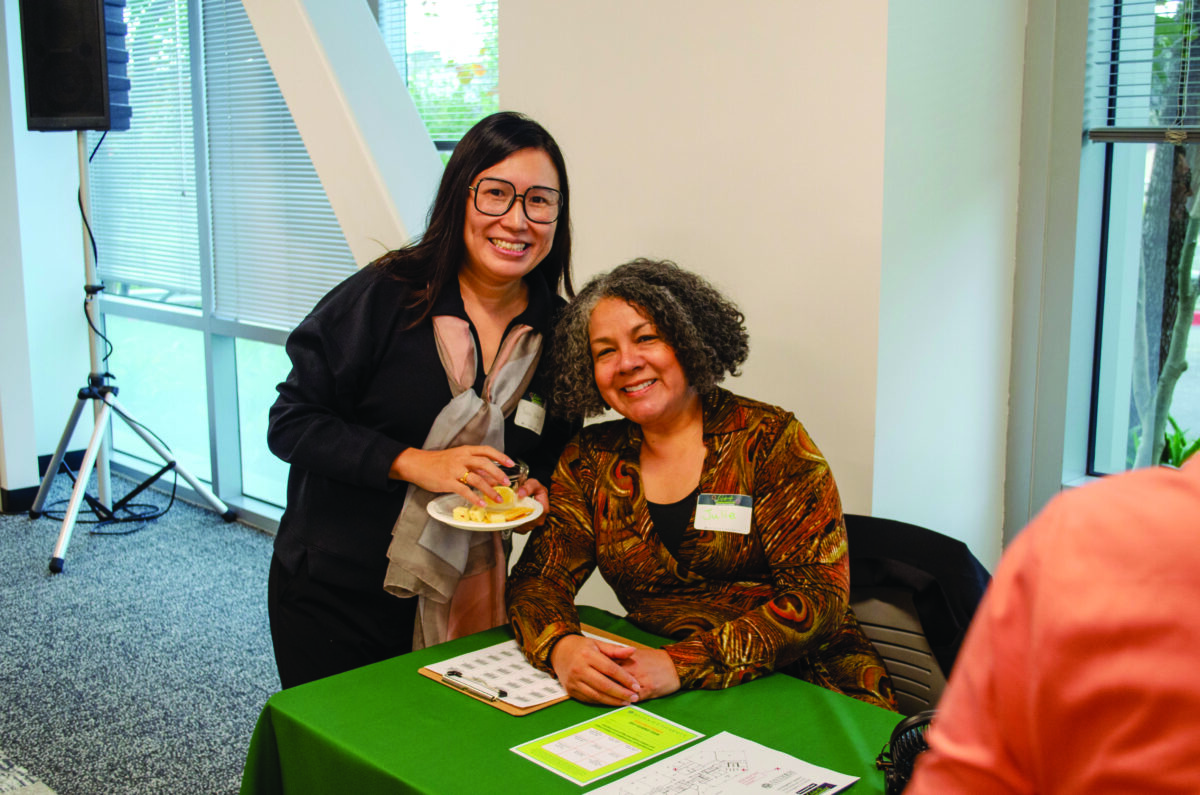 Two women at a table during an event; one stands with a plate of cheese and crackers while the other sits and smiles.