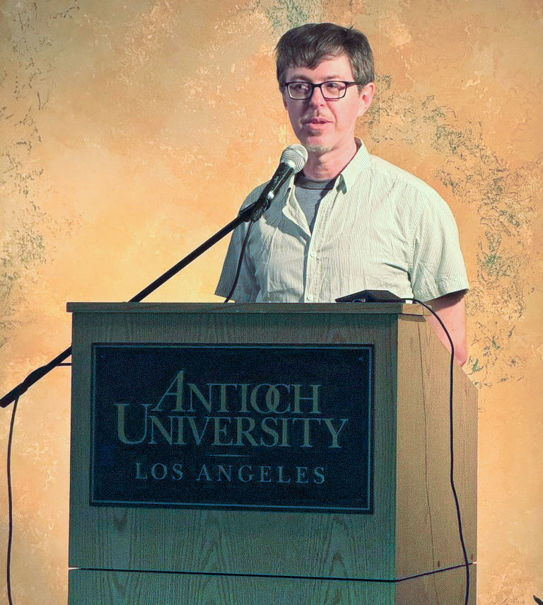Man with glasses speaks into a microphone at a podium labeled Antioch University Los Angeles.