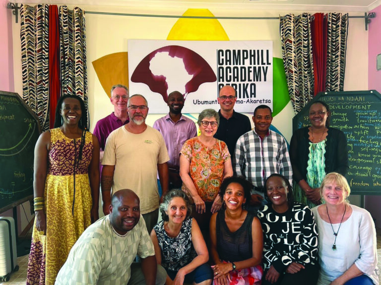Smiling diverse group of adults posing in a bright classroom, with a Camphill Academy Africa banner in the background.