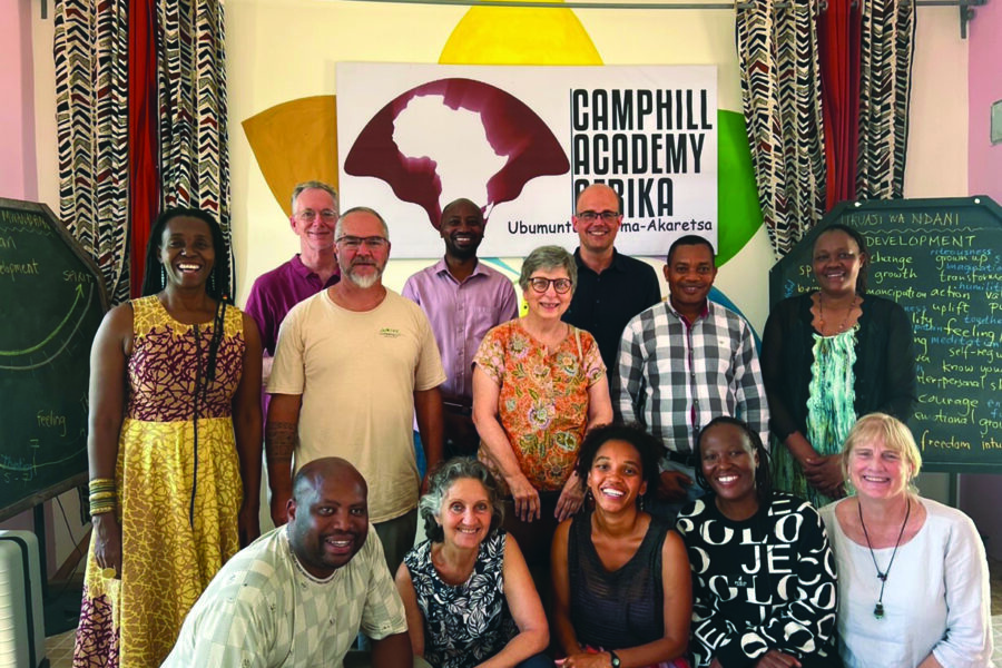 Smiling diverse group of adults posing in a bright classroom, with a Camphill Academy Africa banner in the background.