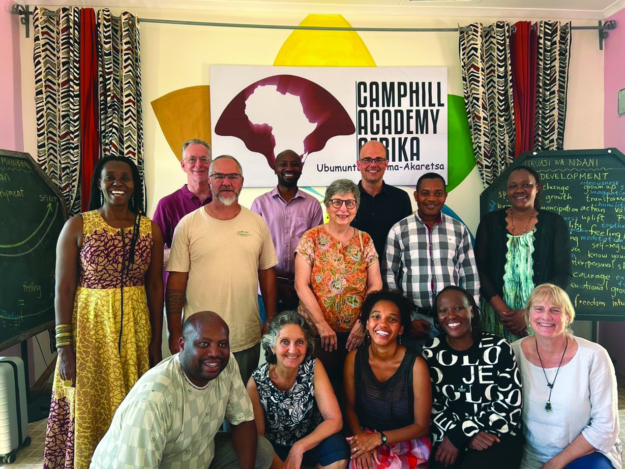 Smiling diverse group of adults posing in a bright classroom, with a Camphill Academy Africa banner in the background.