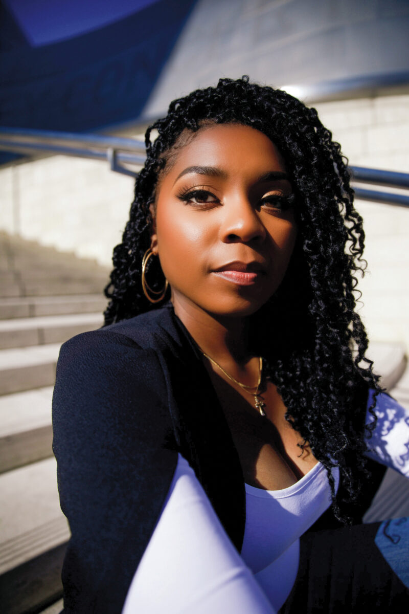 Close-up portrait of a woman with black braided hair, gold hoops, and a navy outer garment outdoors near concrete steps.