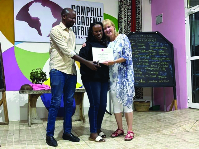 Three adults pose indoors as a certificate is handed to the smiling woman in the center, with a man on the left and another woman on the right.