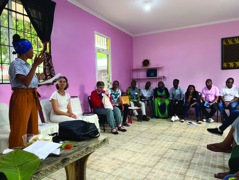 Woman with a blue headwrap and striped top speaks to a group seated in a pink-walled room, addressing the audience with a notebook in hand.