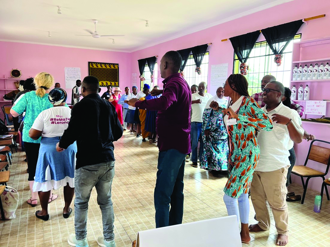 Group of adults dancing in a circle in a bright pink classroom during a community workshop.