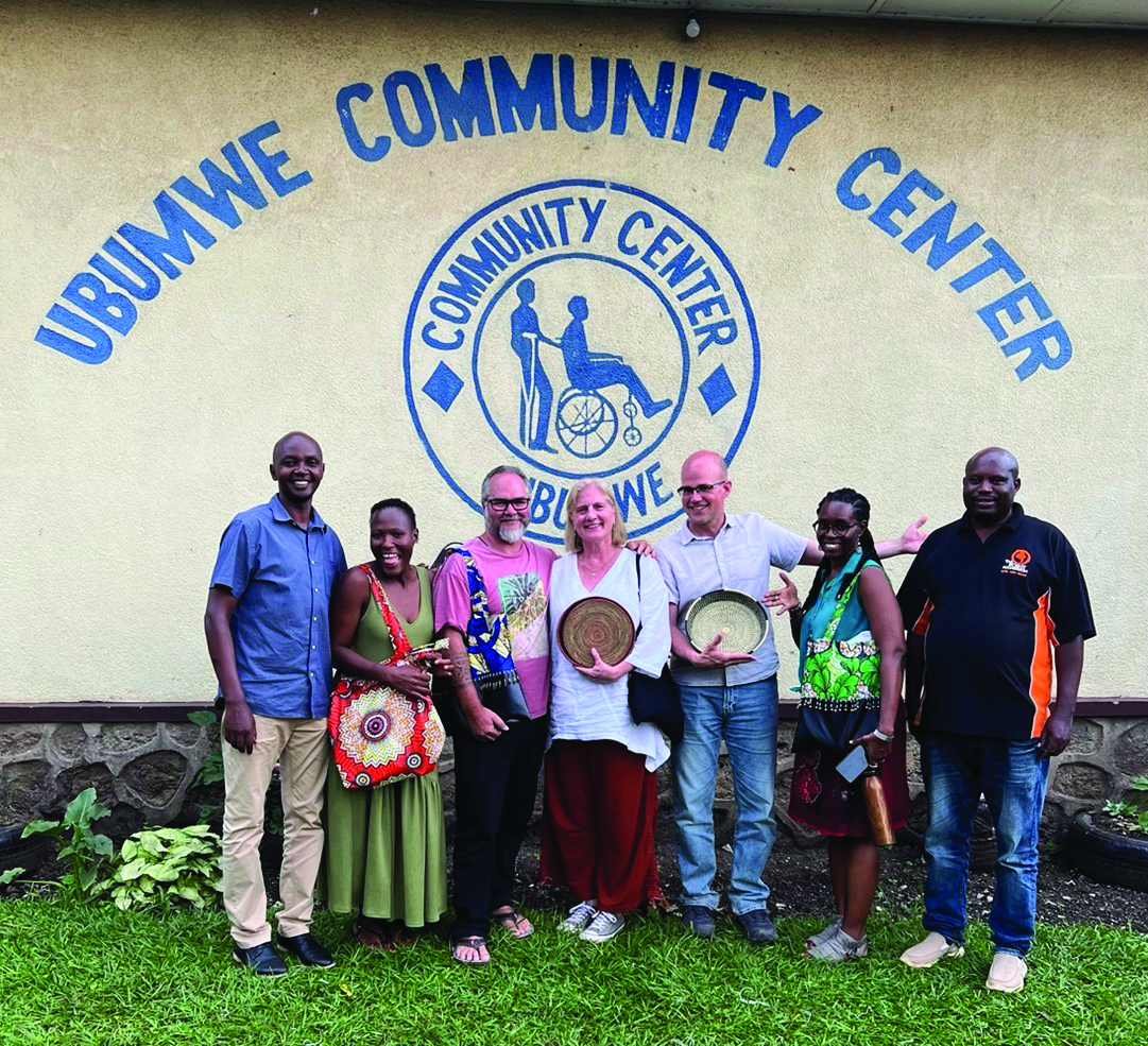 Eight people stand together outside a community center wall, smiling for a group photo; some hold plates and bags.