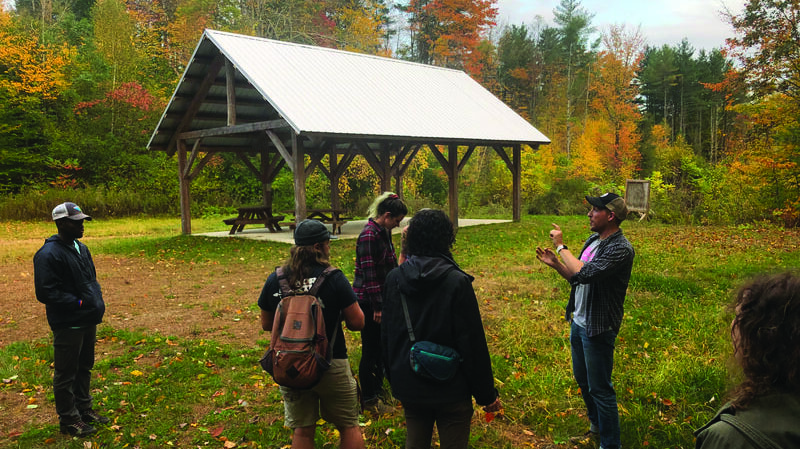 Group of hikers listening to a man explaining something outdoors near a wooden pavilion in a fall forest.