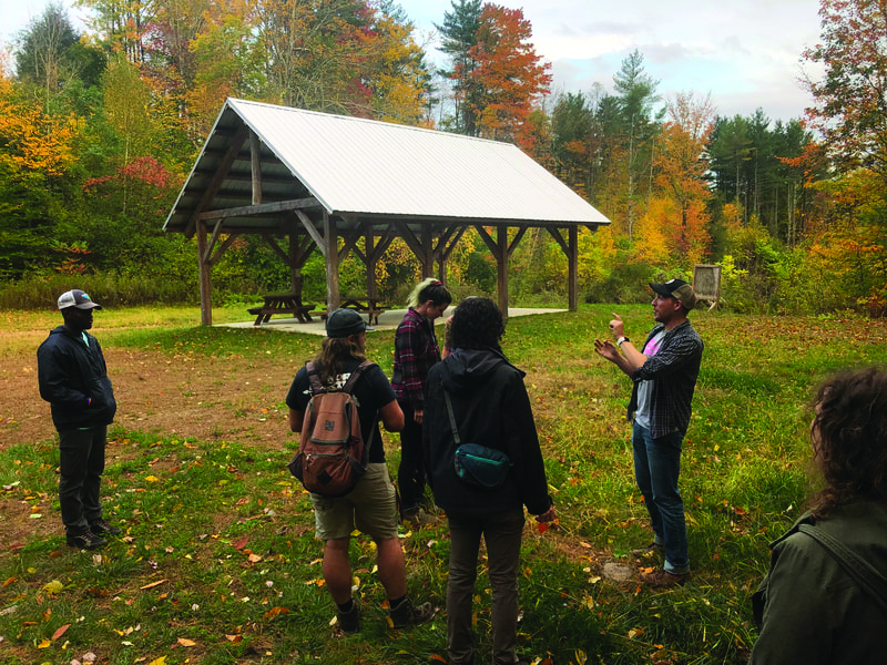 Group of people with backpacks listening to a man speaking near a wooden picnic shelter in autumnal woods.