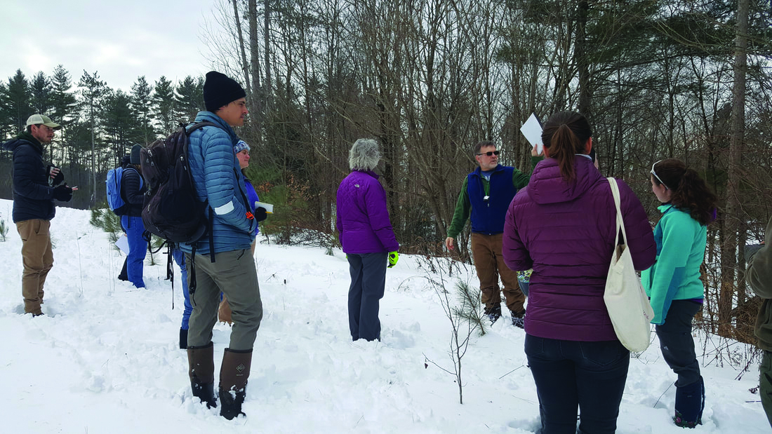 Group of people on a snowy forest hike; guide in a green vest holds up a sheet of paper to the group.