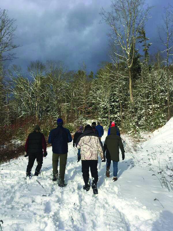 Group of hikers walking through a snow-covered forest trail under a cloudy sky with bare trees.