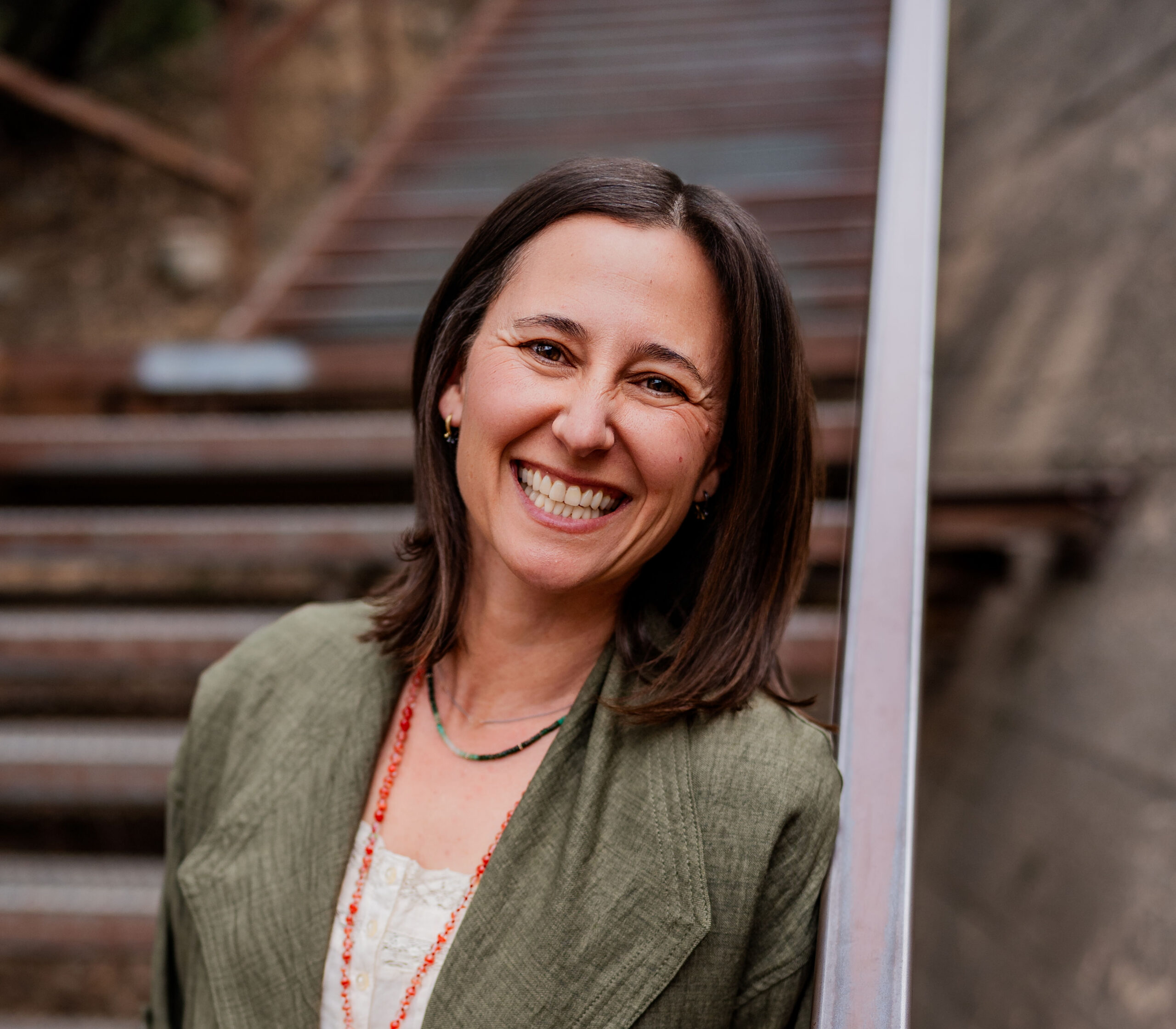 Portrait of a Ileya Grosman smiling. She has shoulder-length dark hair wearing a green blazer over a white top, standing outdoors beside a railing with stone steps in the background.