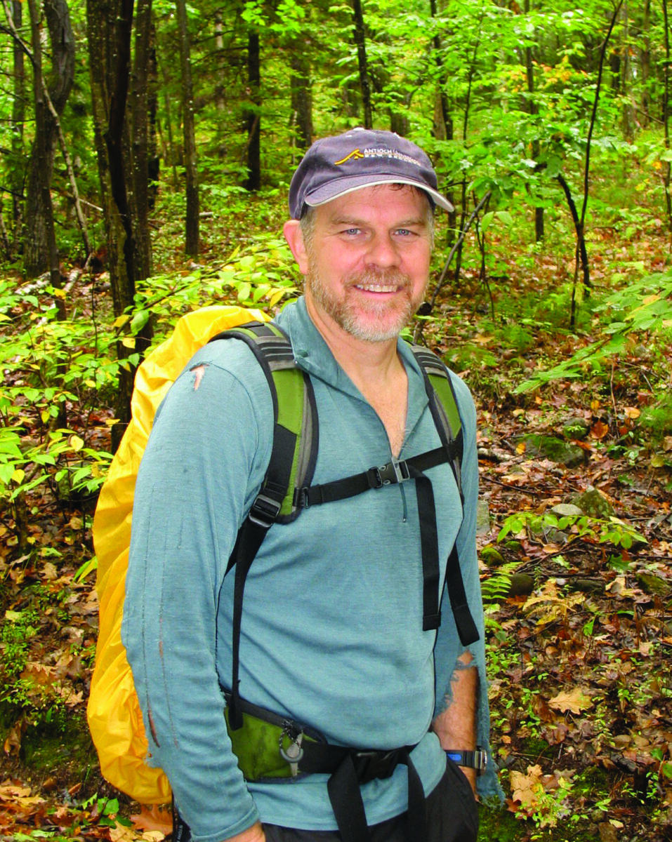Hiker in a green forest wearing a teal long-sleeve shirt and a yellow backpack, smiling at the camera.