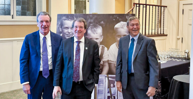 Three men in suits standing together at a formal event in a room with columns and a photo backdrop behind them.