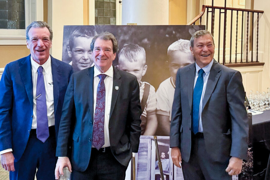 Three men in suits standing together at a formal event in a room with columns and a photo backdrop behind them.
