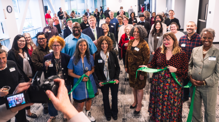 Group of diverse professionals at a ribbon-cutting ceremony in a bright office space, with a photographer in the foreground taking a picture.