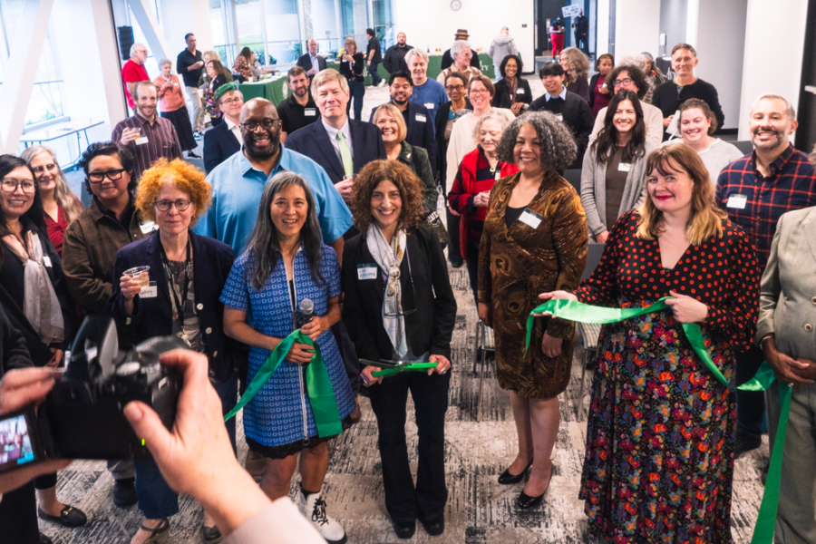 Group of diverse professionals at a ribbon-cutting ceremony in a bright office space, with a photographer in the foreground taking a picture.