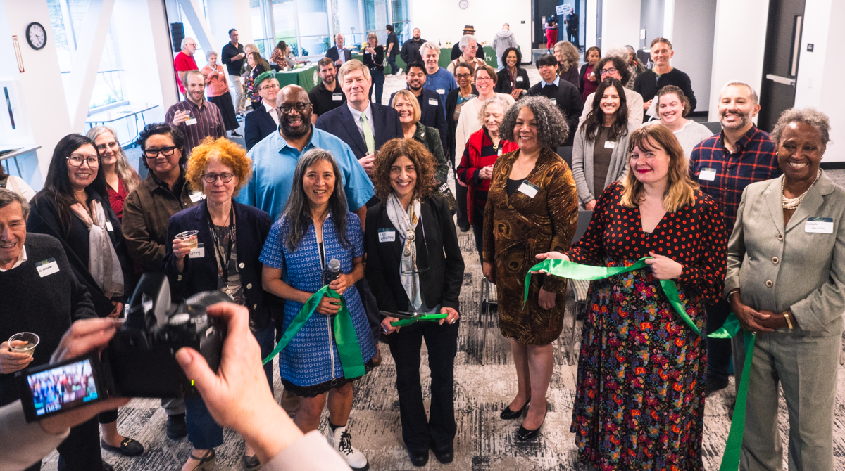 Group of diverse professionals at a ribbon-cutting ceremony in a bright office space, with a photographer in the foreground taking a picture.
