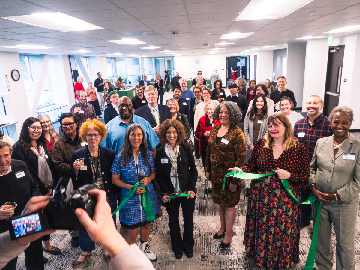 Group of diverse professionals at a ribbon-cutting ceremony in a bright office space, with a photographer in the foreground taking a picture.