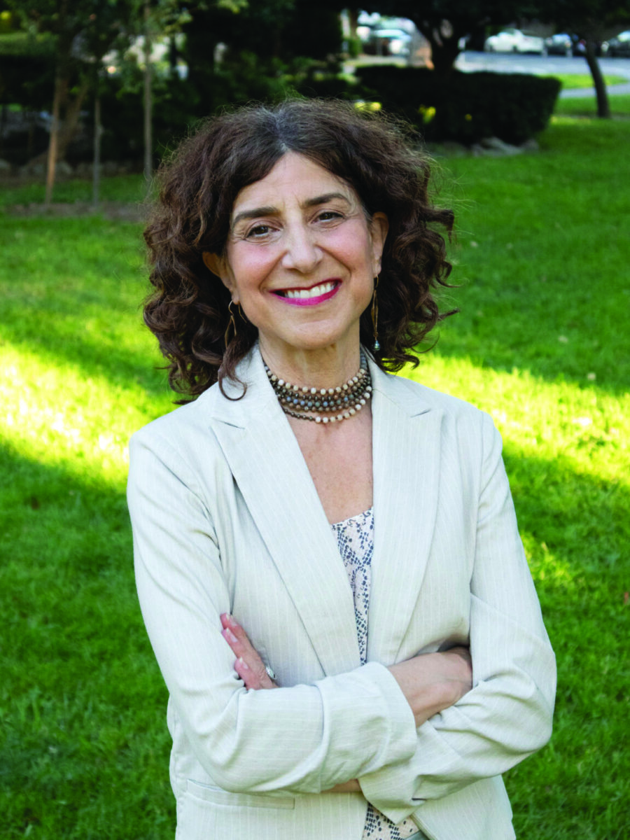 Smiling woman with curly brown hair in a light blazer standing with arms crossed in a sunny park.