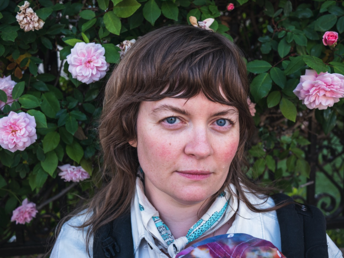Woman with blue eyes and brown hair in front of a bush with pink roses.