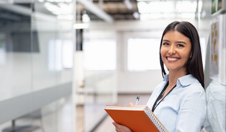 Smiling woman holding an orange notebook and pen in a bright office