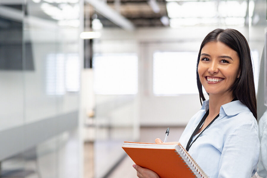 Smiling woman holding an orange notebook and pen in a bright office