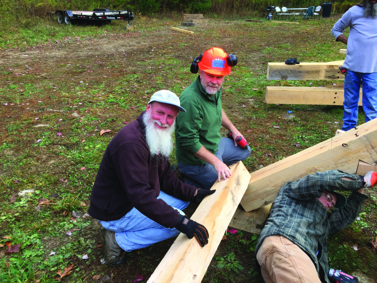 Three men working outdoors on a wooden beam project, with one wearing an orange hard hat and earmuffs, another in a dark cap, and a third partially visible on the right; scattered lumber and construction gloves nearby.