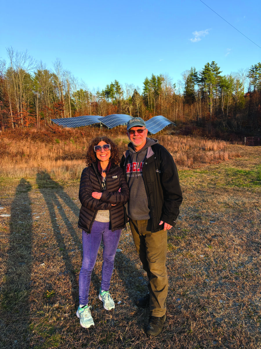 Smiling standing in a grassy field with solar panels in the background and a blue sky above they are wearing jackets.