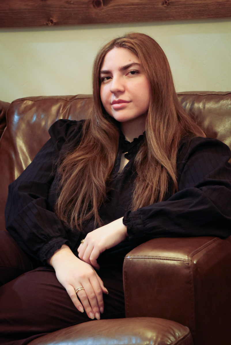 Young woman with long brown hair sits on a brown leather couch, wearing a black blouse, hands resting on her lap, neutral expression.