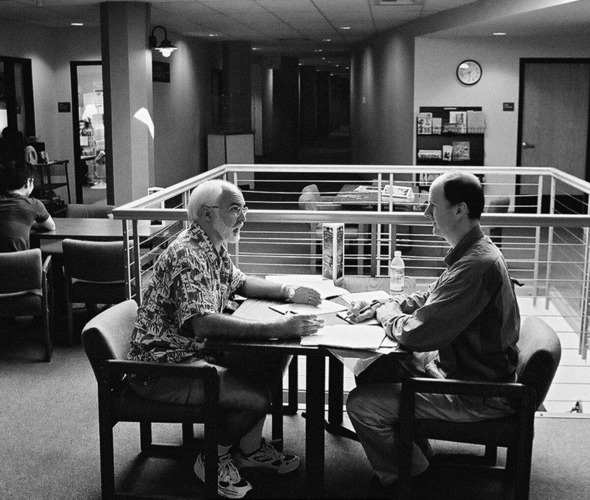 Two men sit at a table in an office, reviewing documents together with pens and a water bottle nearby.