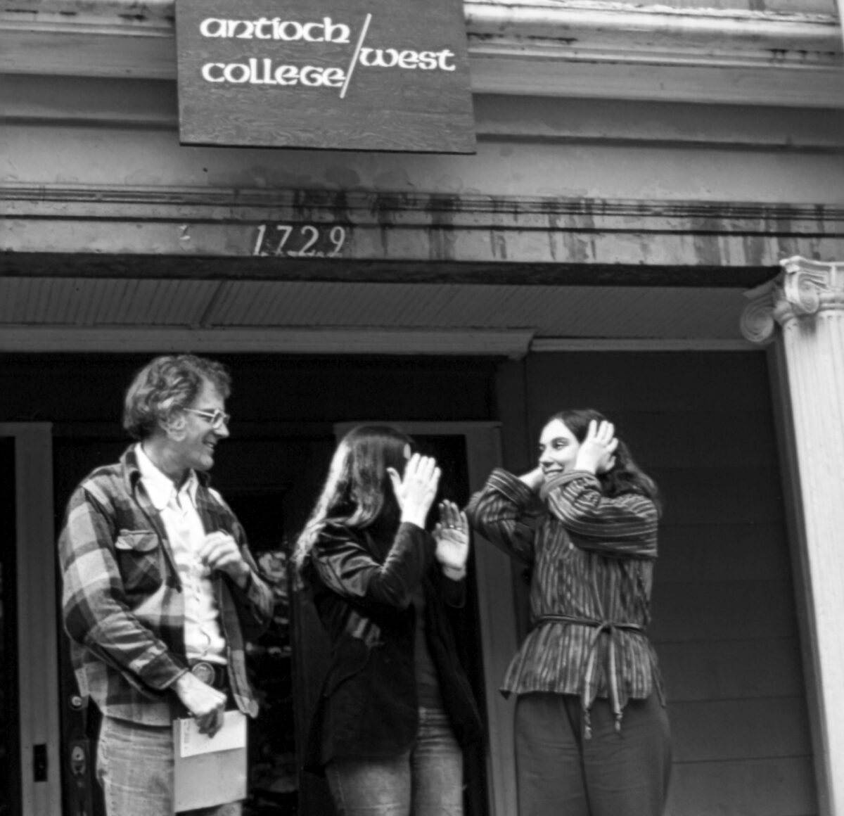 Three adults stand outside a building with a college sign reading 'Antioch College West,' engaging in a lively conversation.